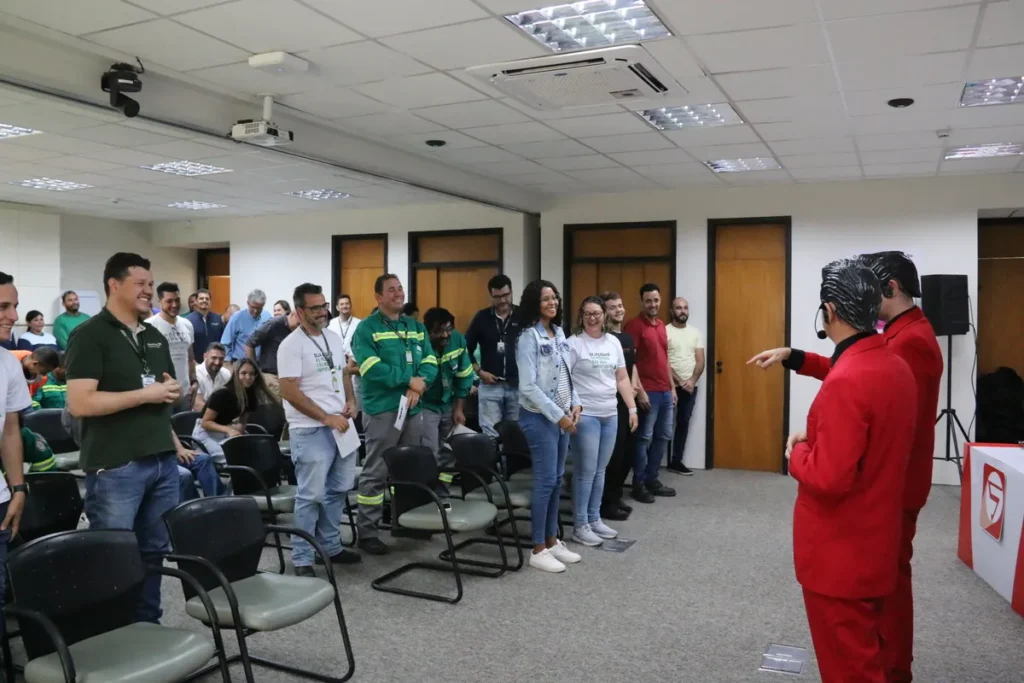 Um grupo de funcionários participa de um evento corporativo, de pé em fila, interagindo com dois palestrantes vestidos de vermelho. A cena representa um treinamento dinâmico sobre ergonomia e segurança no trabalho, com foco no engajamento da equipe.