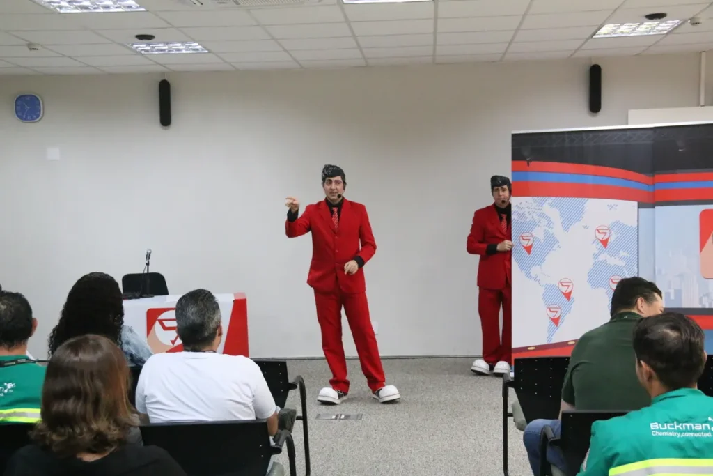 Dois palestrantes fantasiados de terno vermelho ministram um treinamento de ergonomia para um grupo de funcionários, mostrando uma abordagem criativa para conscientização sobre saúde e segurança no trabalho.