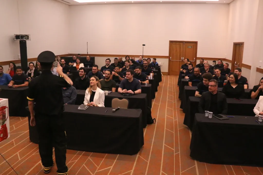 Palestrante de uniforme em frente a uma plateia. Foto de uma reunião de treinamento ou evento corporativo com público sentado em mesas em uma sala de conferências.