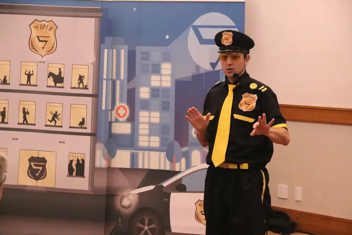 Palestrante de uniforme preto e gravata amarela, com óculos de sol, gesticulando durante uma apresentação em um evento corporativo. O fundo é um painel com tema de super-herói e logo "SUPER 5".