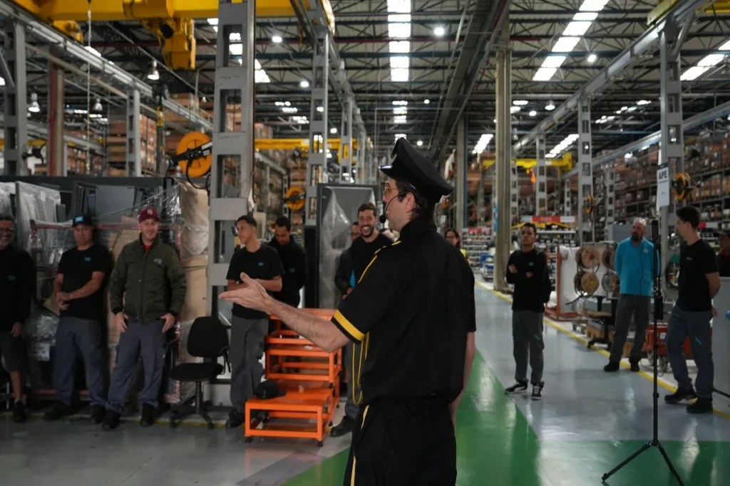 Instrutor de uniforme preto e boné conduzindo uma palestra ou treinamento sobre a NR 01 em ambiente de fábrica ou armazém, com grupo de trabalhadores.