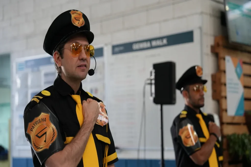 Um homem branco, de óculos de sol laranja e uniforme de segurança preto e amarelo, com a palavra "PATRULHA" escrita na camisa e no boné, gesticula com a mão no peito enquanto fala em um microfone. Atrás dele, um quadro branco com "GEMBA WALK" está visível, e um segundo homem, com uniforme semelhante, está fora de foco.