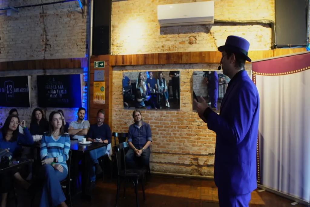 Palestrante sobre alergia alimentar com chapéu e terno azul, falando para a audiência em um local de tijolos aparentes.