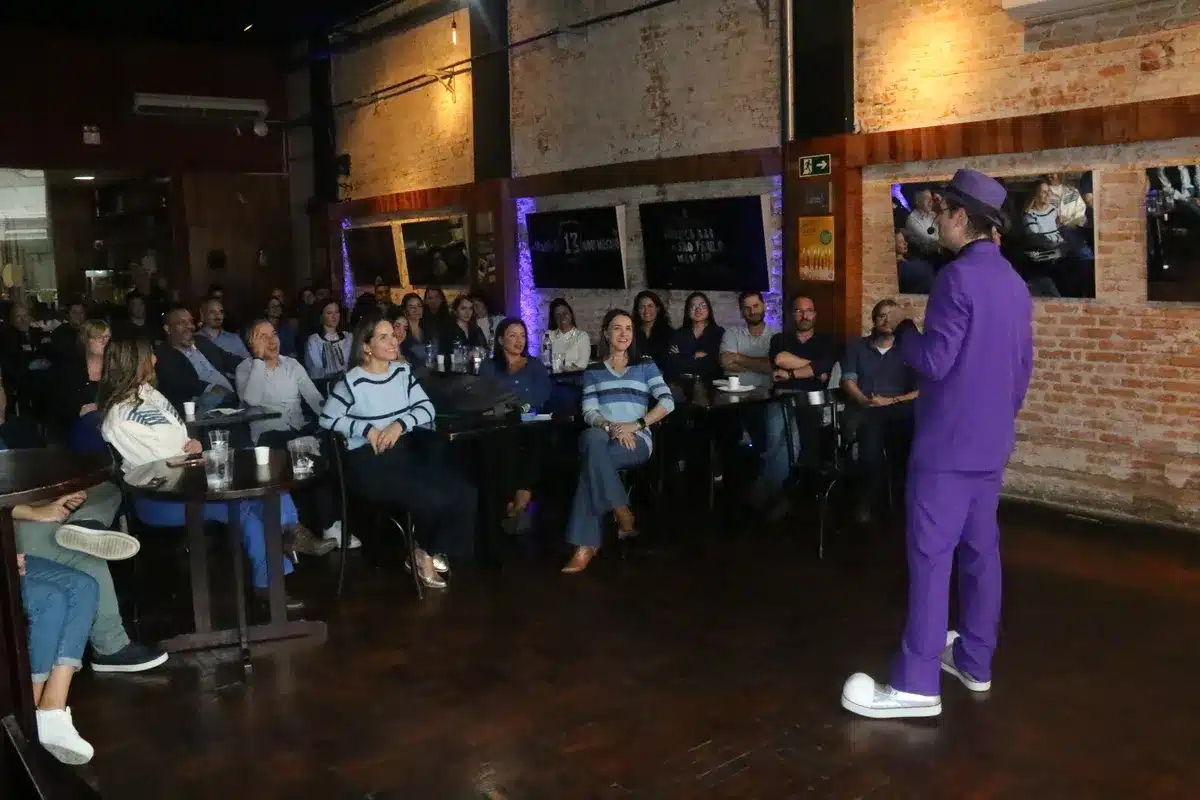Palestrante sobre alergia alimentar com roupa roxa, falando para uma plateia atenta em um bar.