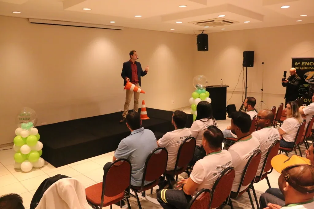 Palestrante em um palco segurando um cone de segurança para um público sentado, simbolizando a importância de sinalização e procedimentos de emergência em treinamentos de primeiros socorros, como a anafilaxia.