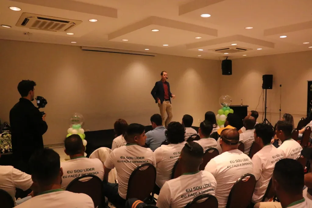 Palestrante masculino caminhando no palco durante um evento de liderança, dirigindo-se a uma plateia que veste camisetas com a frase "Eu sou líder, eu faço a diferença," ilustrando a importância da liderança na resposta a emergências como a anafilaxia.