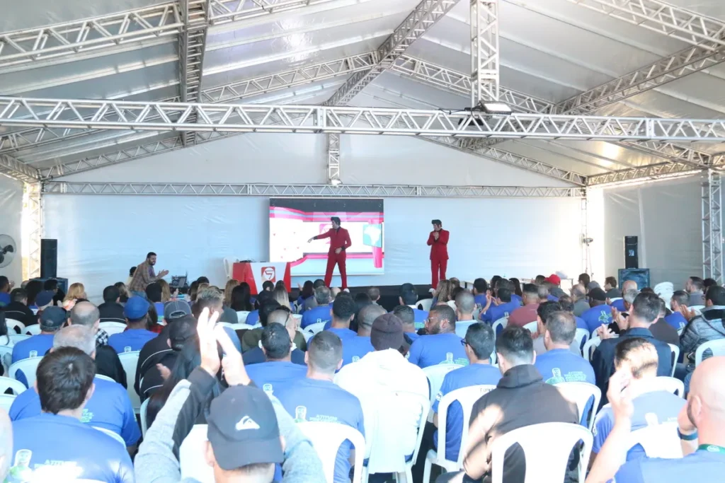 Uma palestra sobre saúde, possivelmente abordando temas como a anemia, com dois palestrantes no palco e uma plateia atenta e engajada. O evento ocorre em uma grande tenda.