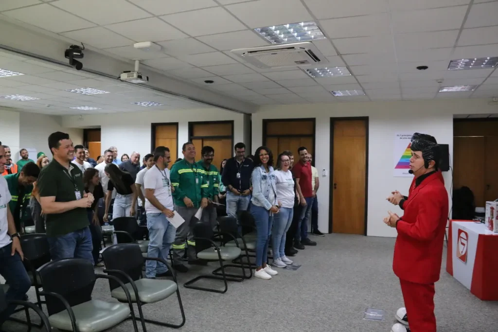 Um palestrante vestido com um terno vermelho e uma peruca preta em estilo Elvis Presley se dirige a um grupo de pessoas em pé, que reagem com sorrisos, em uma sala de conferências.