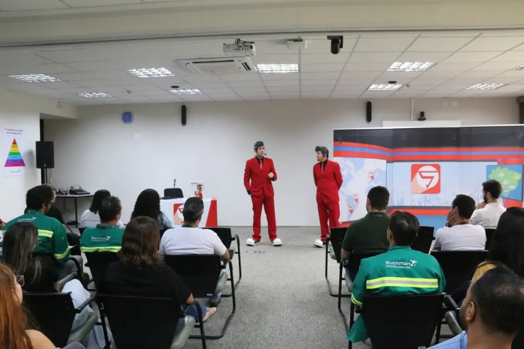 Dois homens, um de terno vermelho e peruca no estilo de Elvis Presley e outro de terno vermelho e boné, estão em um palco se dirigindo a uma plateia em uma sala de conferências. Um telão ao fundo mostra um logotipo e um mapa.