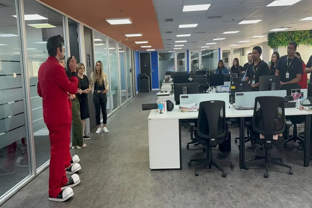 Um homem vestido de forma lúdica, com um terno vermelho e sapatos de palhaço, está de costas, falando com um grupo de pessoas em um escritório. A imagem pode ser usada para ilustrar uma palestra ou workshop sobre saúde e bem-estar, como os cuidados com o assoalho pélvico em ambientes profissionais.
