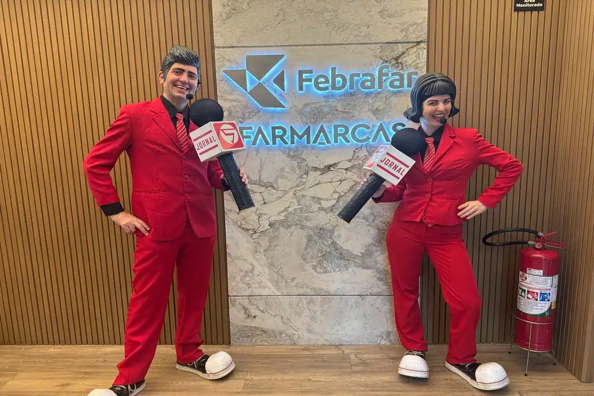 Dois artistas, vestidos com ternos vermelhos e sapatos grandes, seguram microfones com a palavra "JORNAL" e posam em frente aos logotipos das empresas Febralfar e Farmarcas. A imagem lúdica pode ser usada para ilustrar workshops de saúde corporativa, como a importância de exercícios para o assoalho pélvico para o bem-estar dos funcionários.