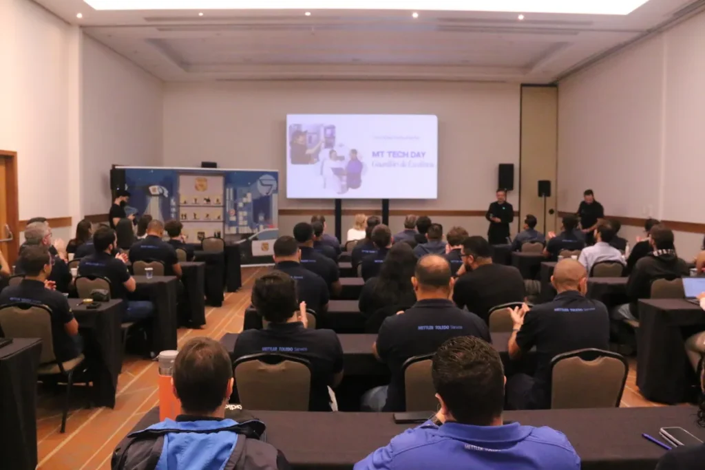 Um auditório lotado com funcionários, a maioria vestindo uniformes escuros da empresa Mettler Toledo Service, assistindo a uma palestra sobre ato inseguro ou segurança técnica. O telão exibe o título "MT TECH DAY – Guardiões da Excelência" com uma imagem de treinamento. A cena representa um evento de conscientização e aprimoramento em segurança e qualidade de trabalho.