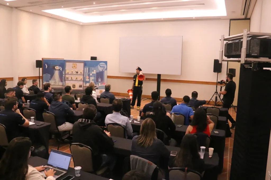 Um palestrante vestido de preto e usando um cone de trânsito como chapéu gesticula à frente de uma tela em branco em uma palestra sobre ato inseguro para um público sentado em mesas escuras. O auditório está cheio de funcionários recebendo treinamento de Segurança e Saúde no Trabalho (SST). A cena representa uma ação de conscientização para evitar falhas humanas e acidentes.