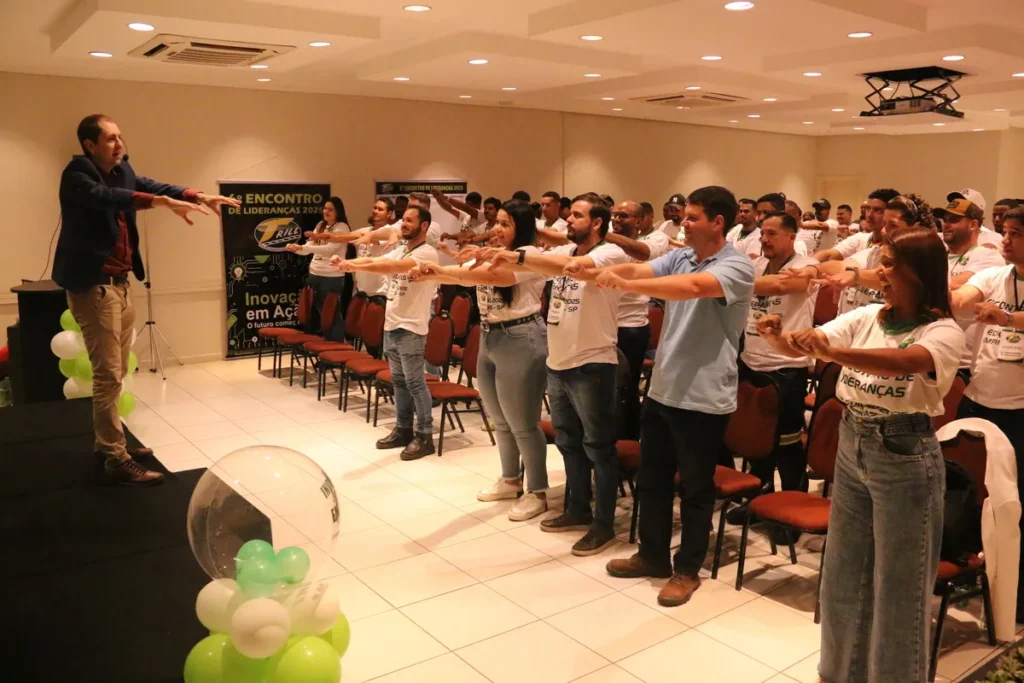 Palestrante de blazer conduz um exercício de alongamento com a plateia em pé, com os braços estendidos, durante uma palestra sobre audiometria e bem-estar.