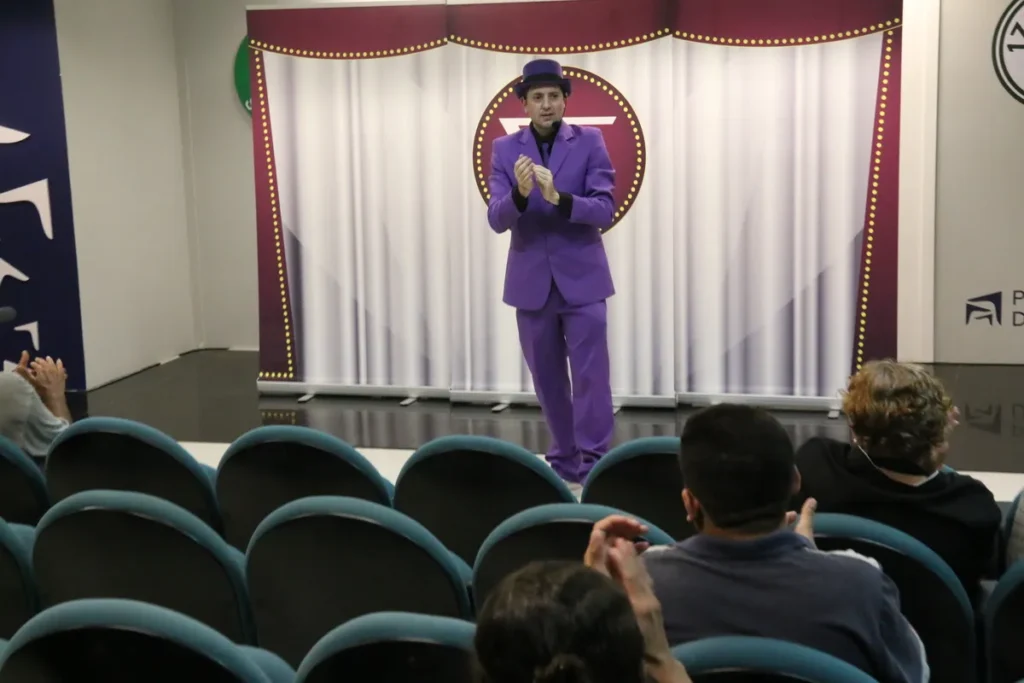 Homem de terno roxo e chapéu apresentando em um palco para uma plateia, com um círculo com um símbolo no fundo, possivelmente relacionado a autismo ou inclusão.