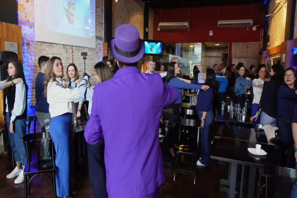 Um instrutor de chapéu e terno roxo, visto de costas, lidera um exercício de alongamento ou dinâmica de grupo com uma audiência em um evento social ou workshop informal.