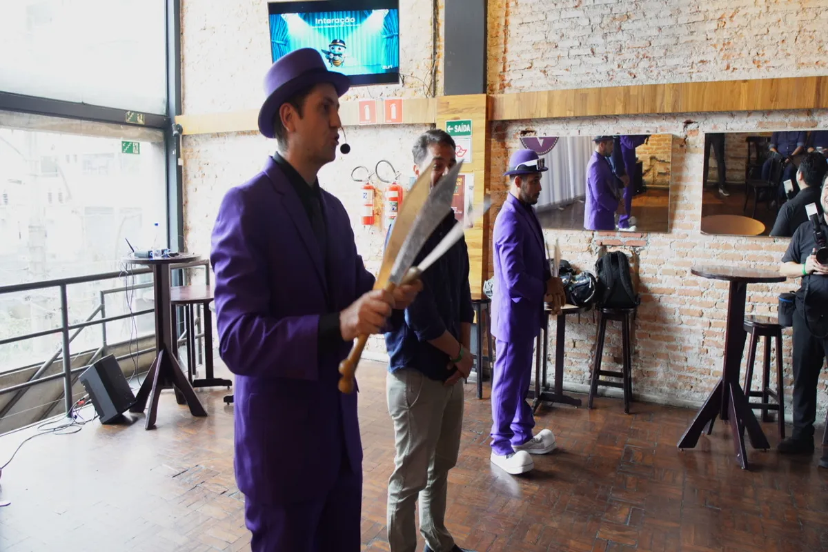 Um homem em terno roxo e chapéu, segurando dois cutelos grandes e gesticulando em um palco, com outro homem de terno roxo ao lado, em um evento informal.