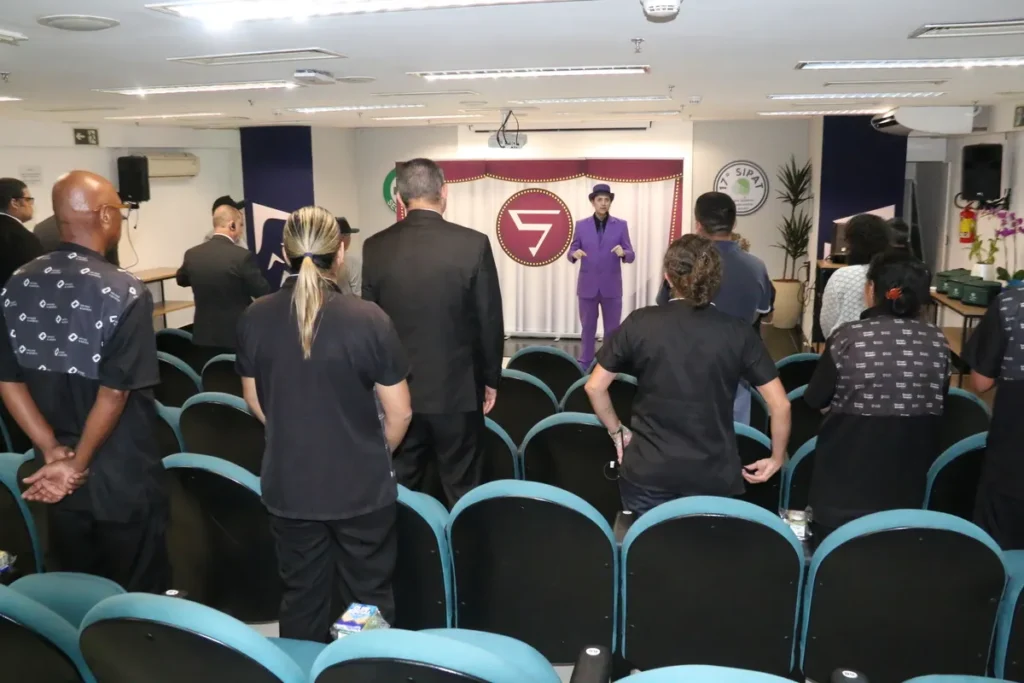 Público de costas assistindo a um palestrante de terno roxo e chapéu em um palco, com um logotipo ao fundo que pode representar uma organização de saúde mental ou evento.