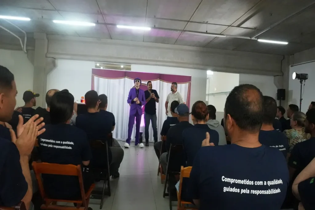 Um grupo de pessoas está assistindo a uma palestra. Um palestrante, vestido com um terno roxo, e um homem com uma camisa polo estão em um palco. A imagem sugere um evento sobre conscientização de temas como o bullying.