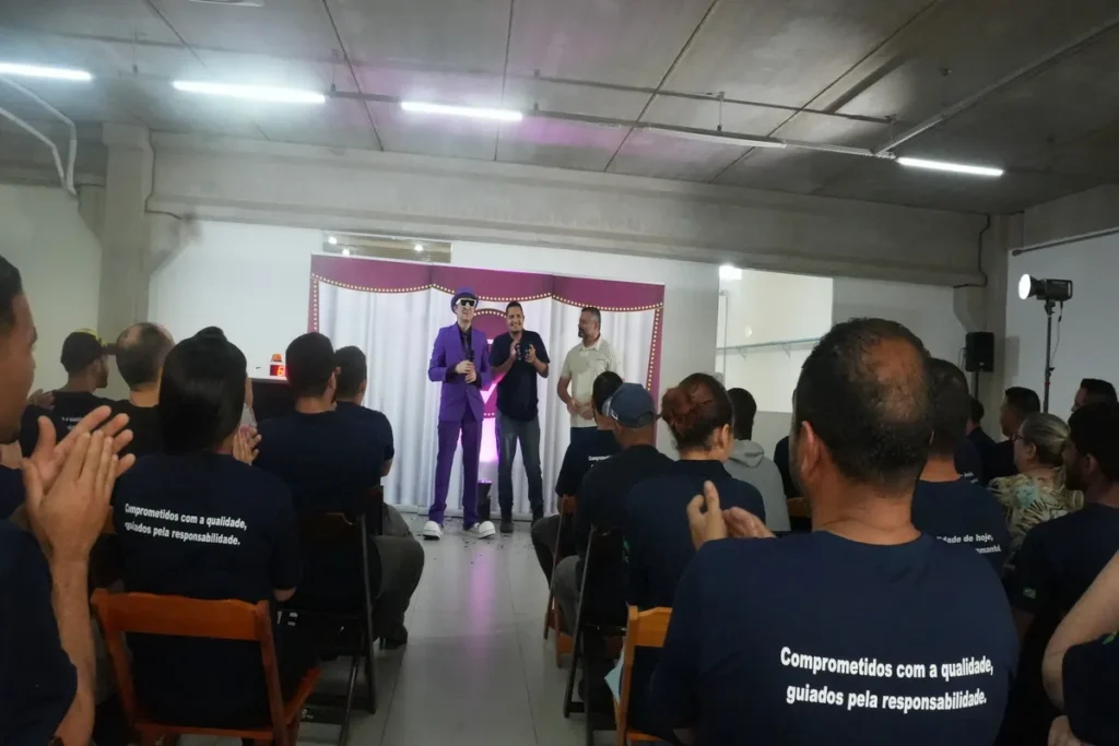 Um grupo de pessoas está assistindo a uma palestra. Um palestrante, vestido com um terno roxo, e um homem com uma camisa polo estão em um palco. A imagem sugere um evento sobre conscientização de temas como o bullying.