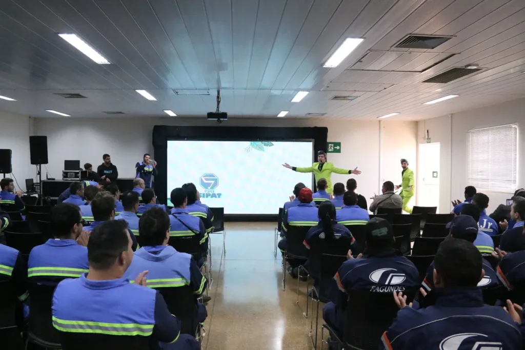 Dois palestrantes em ternos verde-limão interagem no palco de um auditório, em frente a uma tela grande exibindo o logo "SUPER SIPAT". Eles estão fazendo uma apresentação para uma plateia de funcionários vestidos com uniformes da empresa (com o logo Eurochem nas costas). A cena representa uma palestra de Saúde e Segurança do Trabalho (SST), crucial para a prevenção de doenças como a bursite.