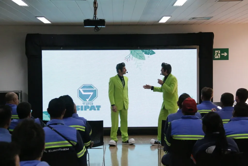 Dois palestrantes vestidos com ternos verde-limão e sapatos brancos fazendo uma apresentação animada para um público de funcionários (vestidos com uniformes azuis e amarelos da Eurochem) em um auditório. O telão ao fundo exibe o logotipo "SUPER SIPAT". A cena é de uma palestra sobre saúde e segurança do trabalho, ideal para temas como a prevenção de bursite e outras lesões ergonômicas.