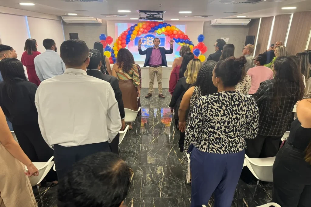 Um palestrante se dirige a um público diverso de funcionários em um evento corporativo, decorado com um grande arco de balões coloridos (vermelho, amarelo, azul) e o logo "GB" no telão. A palestra ocorre em um espaço interno e bem iluminado. A cena representa um momento de conscientização e engajamento da equipe durante a Campanha Outubro Rosa.