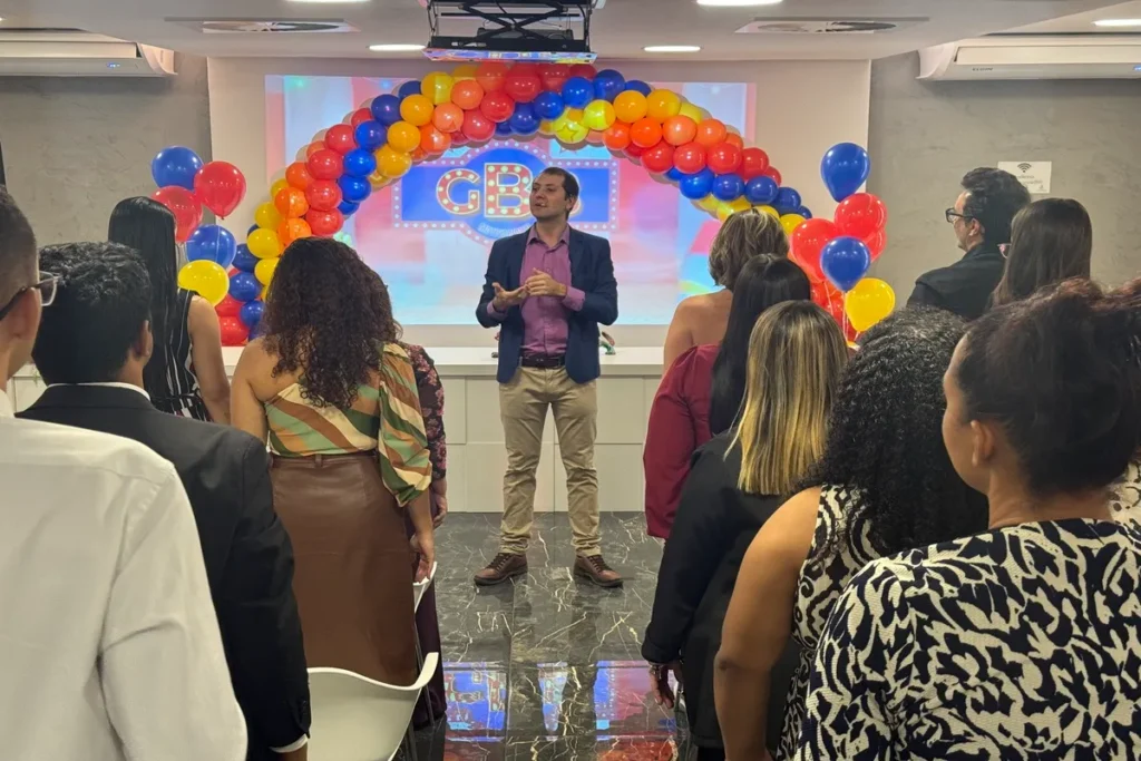 Um palestrante (homem, com blazer azul) se dirige a um público de funcionários (principalmente mulheres) em uma sala decorada com um arco de balões coloridos (vermelho, amarelo, azul), indicando um evento corporativo e festivo, como o lançamento da Campanha Outubro Rosa de conscientização sobre o câncer de mama.