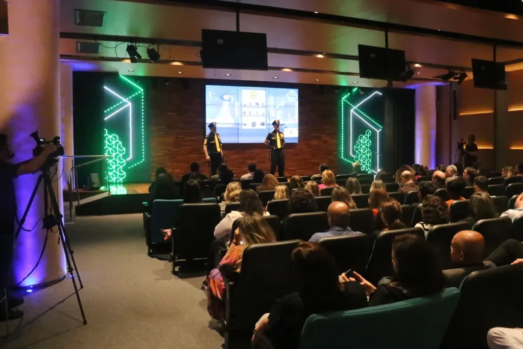 Dois homens em trajes de policial de desenho animado, com camisas pretas e amarelas, se apresentam em um palco com iluminação neon verde e um telão ao fundo. Uma plateia assiste sentada em fileiras de cadeiras em um auditório.