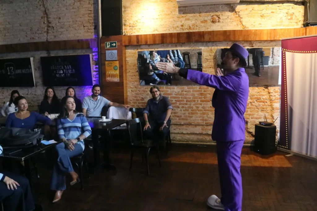 Palestrante com terno e chapéu roxo gesticulando para a plateia durante uma apresentação em um ambiente com paredes de tijolo.
