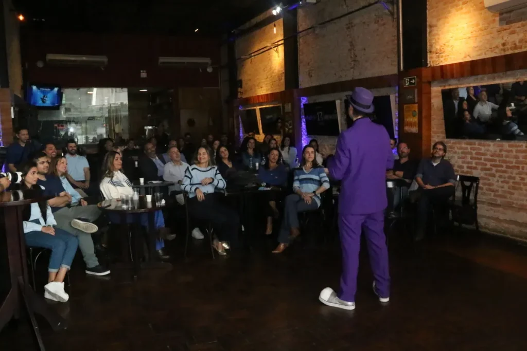 Palestrante de terno e chapéu roxo se apresenta para uma plateia sentada em um evento noturno com iluminação de palco.