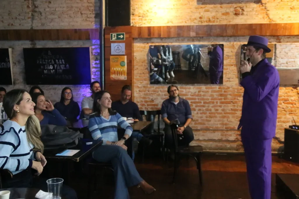 Palestrante com terno e chapéu roxo falando para a plateia que está sorrindo e assistindo em um evento noturno.