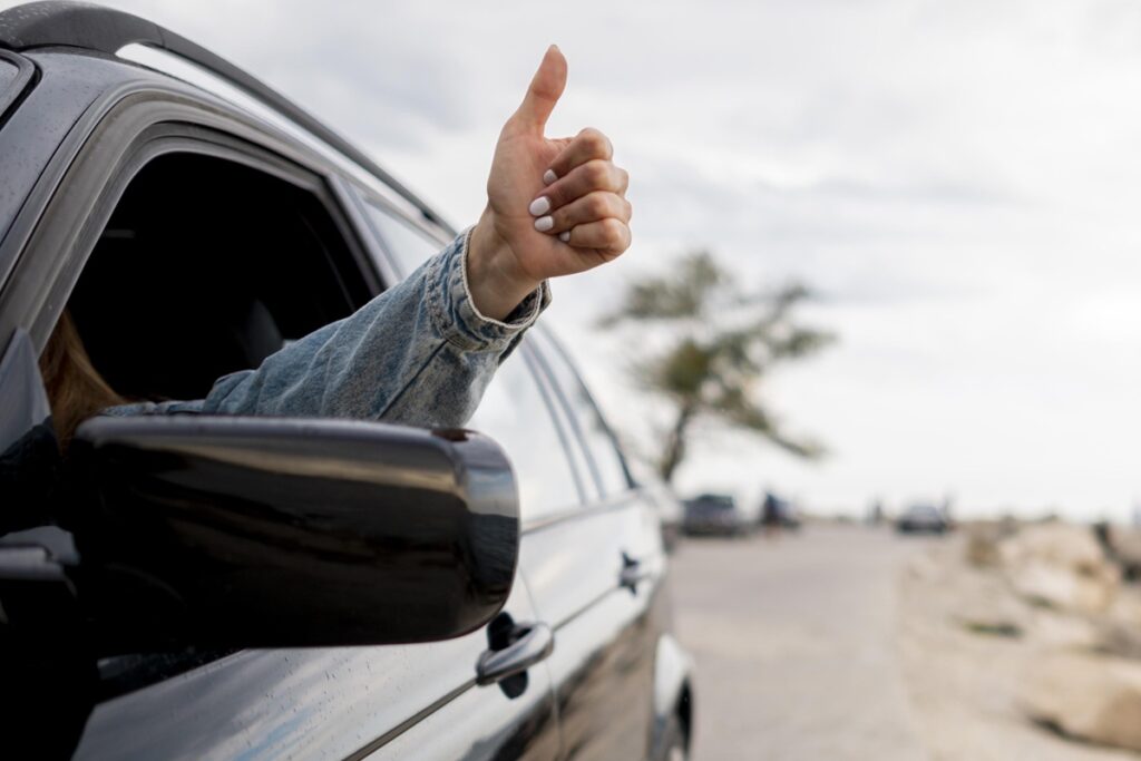 Braço de motorista para fora da janela fazendo sinal de positivo, indicando oferta de carona ou aprovação durante o trajeto.