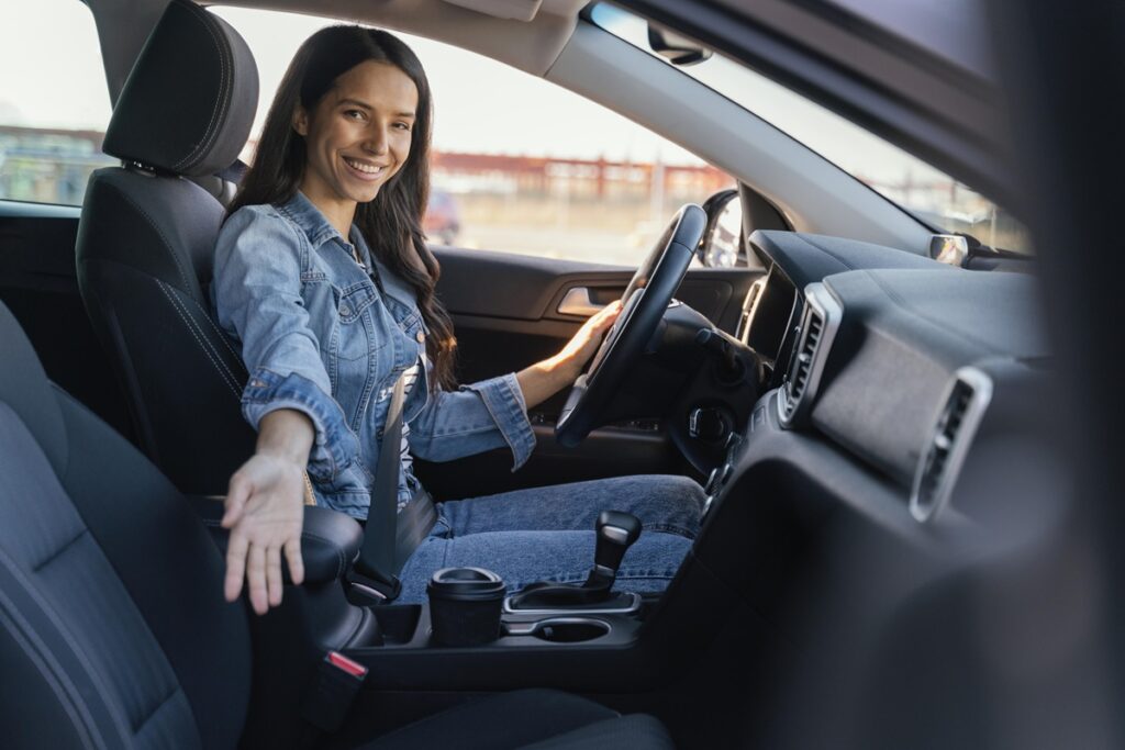 Mulher ao volante sorrindo e estendendo a mão para convidar alguém a entrar no carro, representando carona solidária.