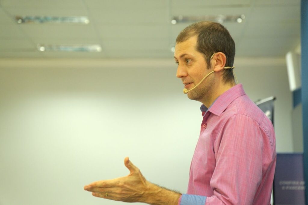 Palestrante usando microfone headset e camisa rosa durante apresentação corporativa de SIPAT.