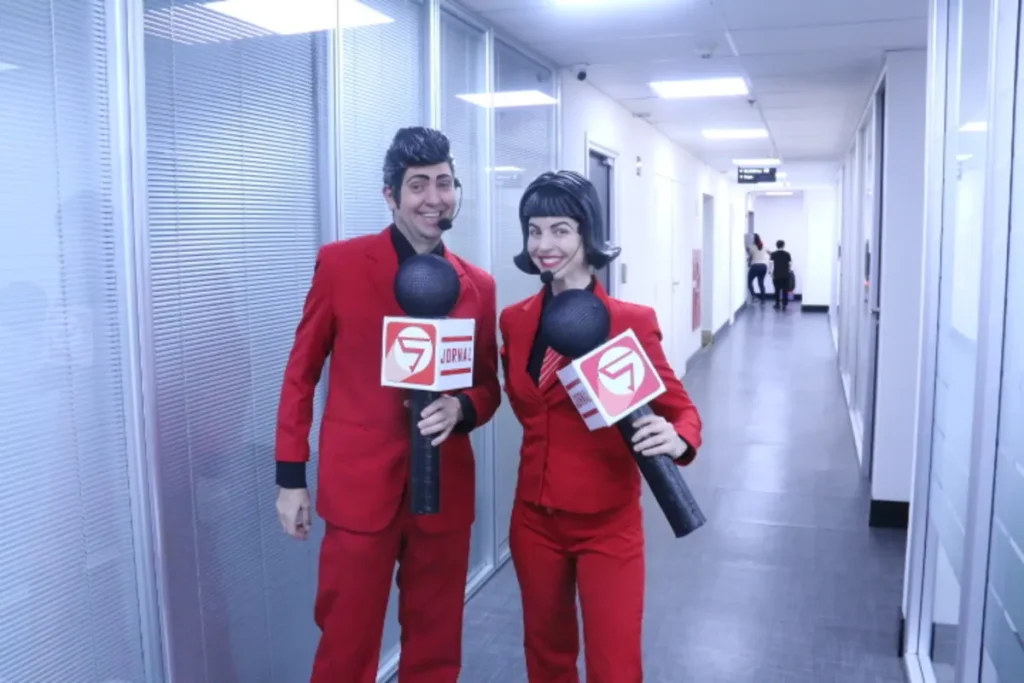 Um homem e uma mulher, vestidos com ternos vermelhos brilhantes e perucas estilizadas, seguram microfones com o logo "Jornal 7" em um corredor de escritório. Eles representam personagens de jornalistas em uma ação de conscientização da CIPA ou SIPAT, buscando comunicar temas de segurança no trabalho de forma criativa.