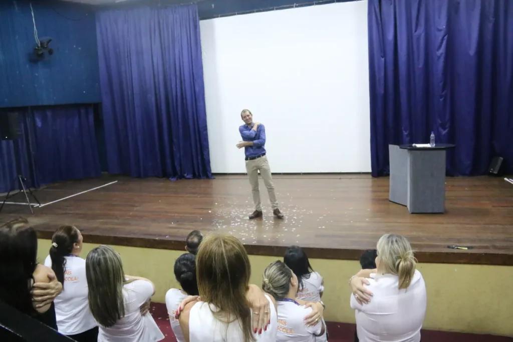 Homem de pé em um palco, com os braços cruzados, em uma palestra de coaching motivacional para uma plateia.