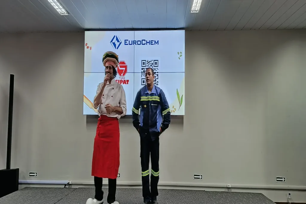 Palestrante vestido de chef de cozinha e um funcionário com uniforme de trabalho no palco, apresentando uma palestra sobre compulsão alimentar em frente a um telão da EuroChem.