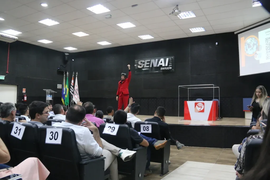 Palestrante de terno vermelho no palco, erguendo o punho para uma plateia atenta em um auditório do SENAI, em uma palestra sobre compulsão alimentar.