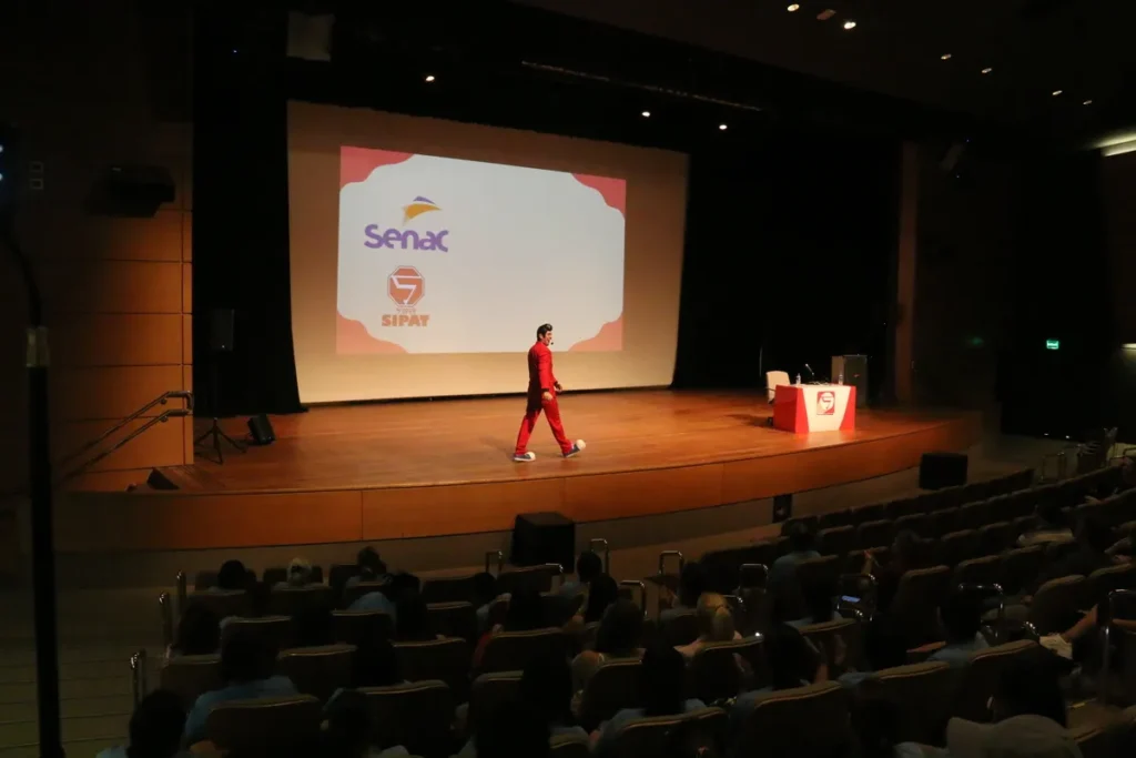 Um homem vestido com terno vermelho e em estilo de personagem (Cipeiro) em um palco de auditório, chutando uma bola de futebol em direção à plateia, enquanto um telão exibe os logotipos do SENAC e SIPAT (Semana Interna de Prevenção de Acidentes de Trabalho).