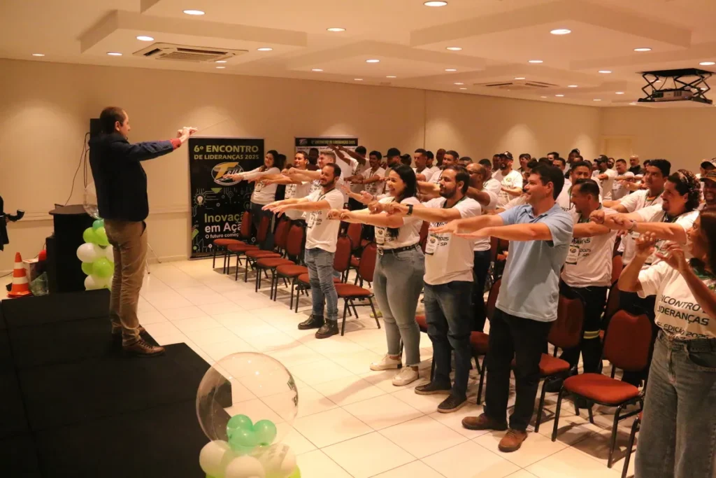 Um palestrante de terno está em um palco, conduzindo uma dinâmica de alongamento ou interação com uma grande plateia de colaboradores. Os participantes, vestindo camisetas brancas do "6º Encontro de Liderança", estão em pé com os braços esticados para os lados, seguindo a instrução. Este momento de aquecimento é comum em treinamentos de segurança e pode ser adaptado para o formato DDS online, reforçando o engajamento e a importância do Diálogo Diário de Segurança.