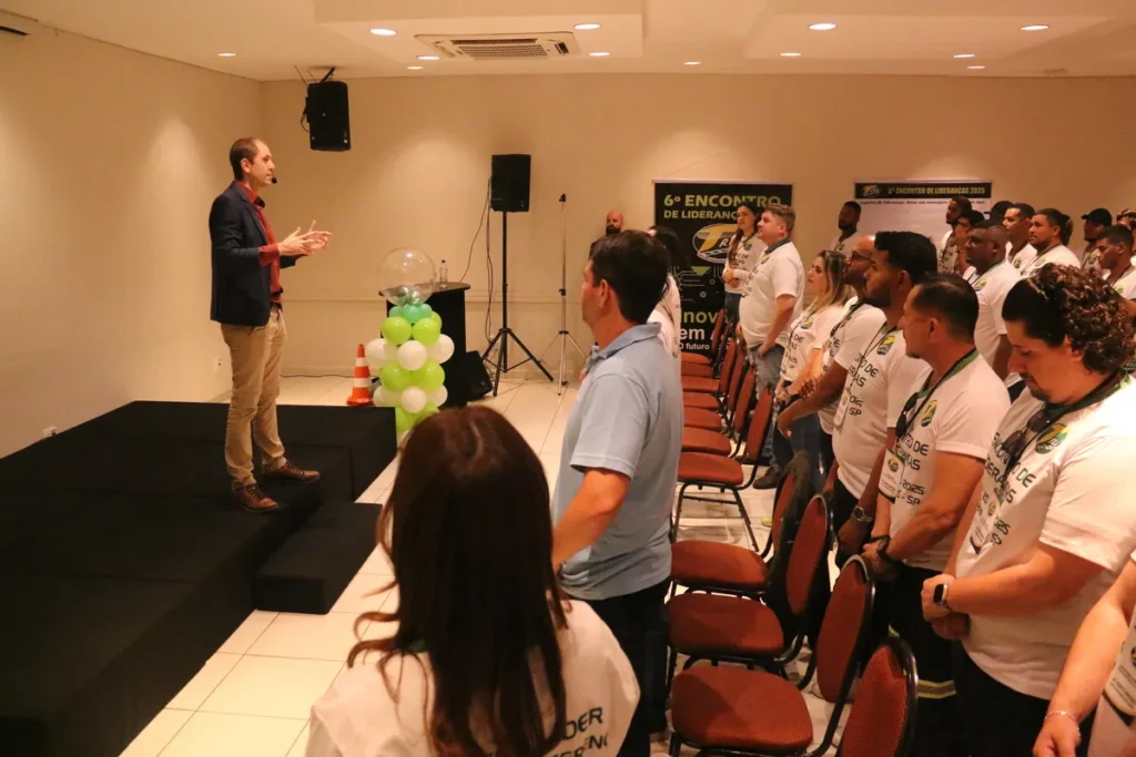 Um palestrante de terno e calça cáqui discursa em um pequeno palco elevado, gesticulando para uma plateia de profissionais. A plateia está de pé e atenta, vestindo camisetas brancas de um "6º Encontro de Liderança". O tema do evento remete a segurança e treinamento. Há um totem decorativo com balões verdes e brancos. A palavra-chave DDS online pode ser associada ao contexto de treinamentos e Diálogos Diários de Segurança.