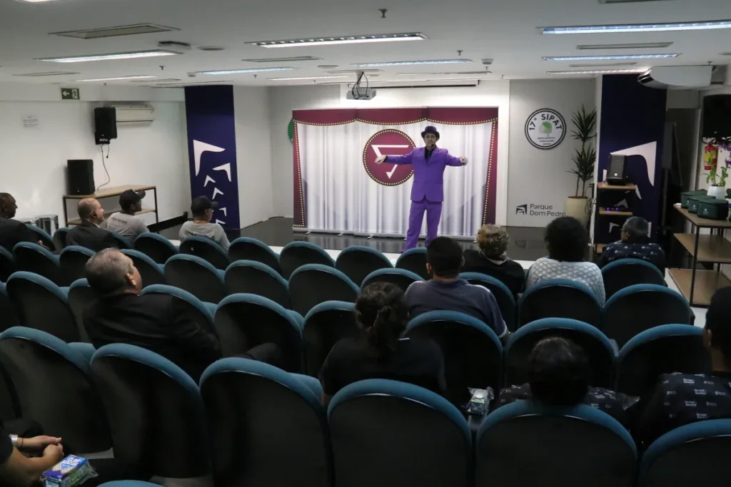 Um palestrante, em um terno roxo vibrante e um chapéu, fala para uma plateia em um auditório. A imagem simboliza a conscientização sobre dislexia e outros temas.