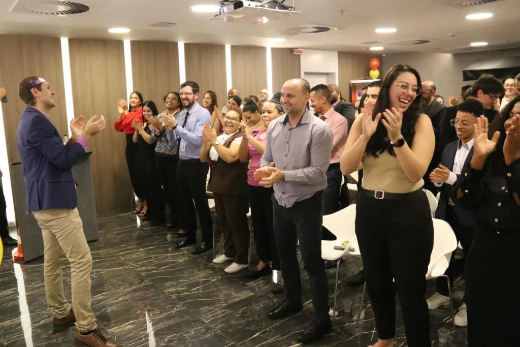 Pessoas em um evento de saúde, incluindo profissionais e público, aplaudem de pé um palestrante em um auditório moderno, com o tema relacionado à 'doença arterial coronariana' ou saúde cardiovascular.