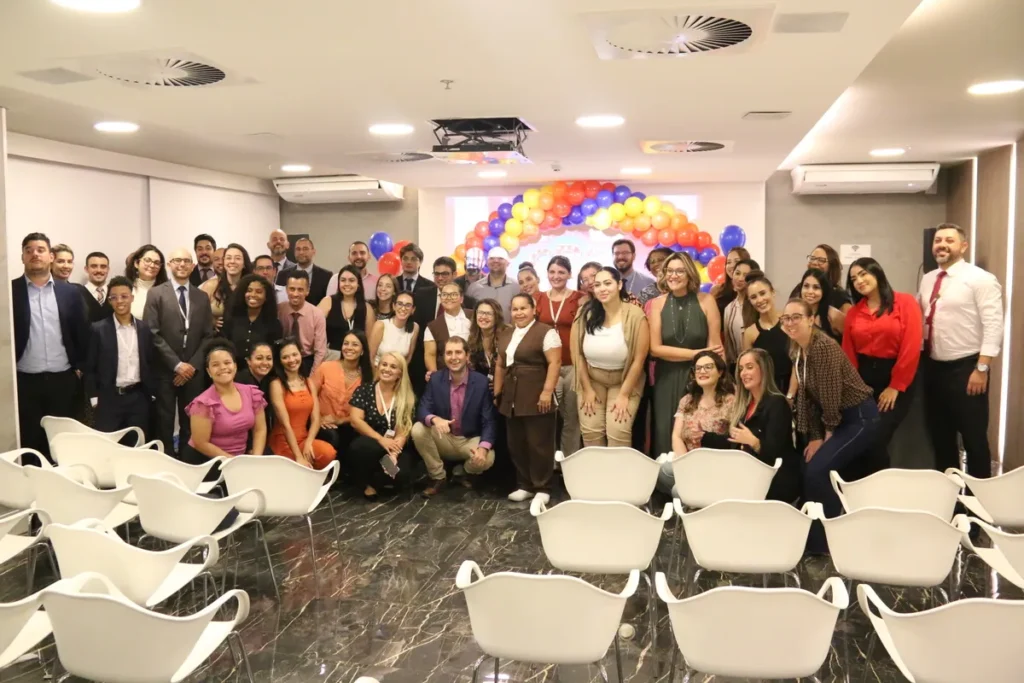 Um grande grupo de pessoas, incluindo homens e mulheres, posando para uma foto em um evento de saúde corporativa, com um arco de balões ao fundo, ilustrando uma campanha de conscientização sobre 'doença arterial coronariana'.