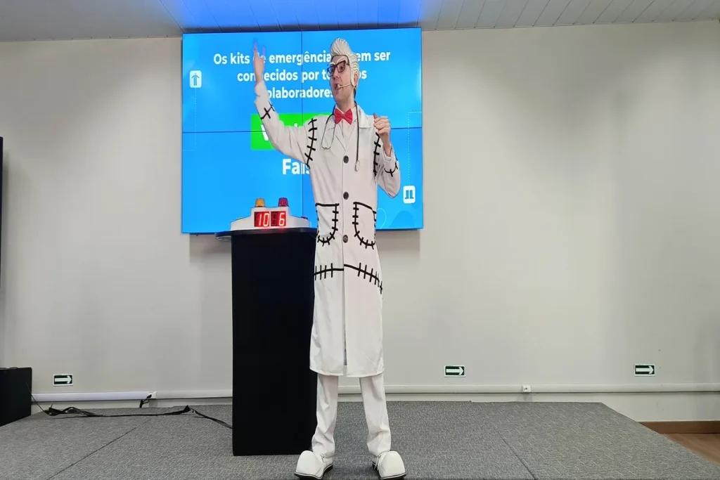 Homem fantasiado de "Dr. Dor Lombar" em um palco, com um jaleco branco costurado e maquiagem facial de pontos, fazendo uma apresentação em frente a um telão com a frase "Os kits de emergência devem ser conhecidos por todos os colaboradores".
