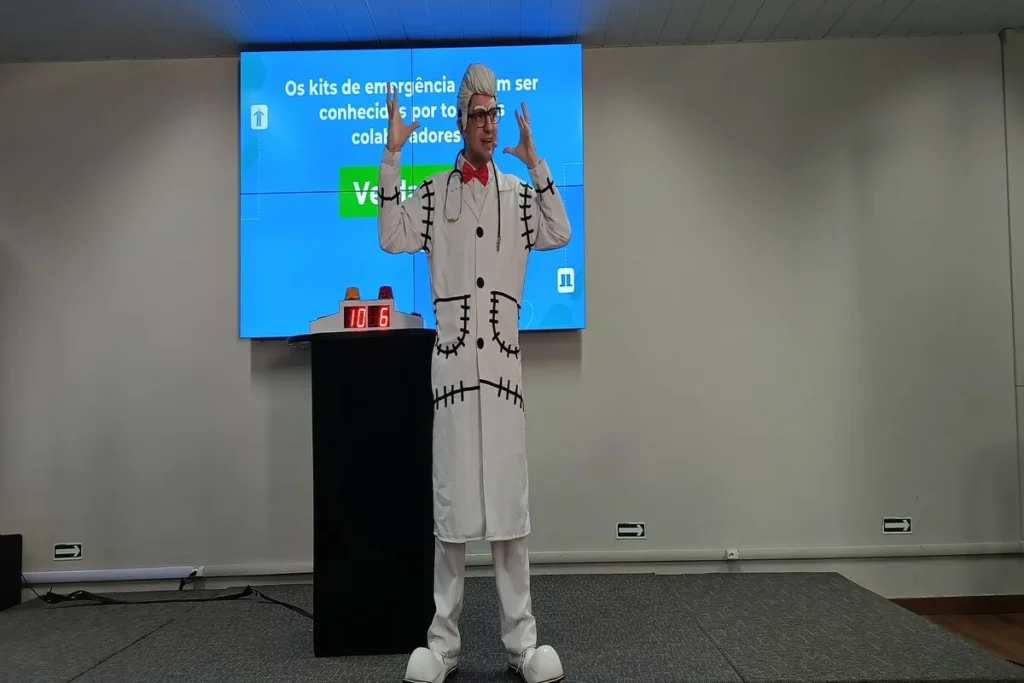 Homem fantasiado com jaleco branco com pontos pretos e maquiagem facial, fazendo uma apresentação sobre dor lombar crônica em frente a um telão azul com a frase "Os kits de emergência devem ser conhecidos por todos os colaboradores".