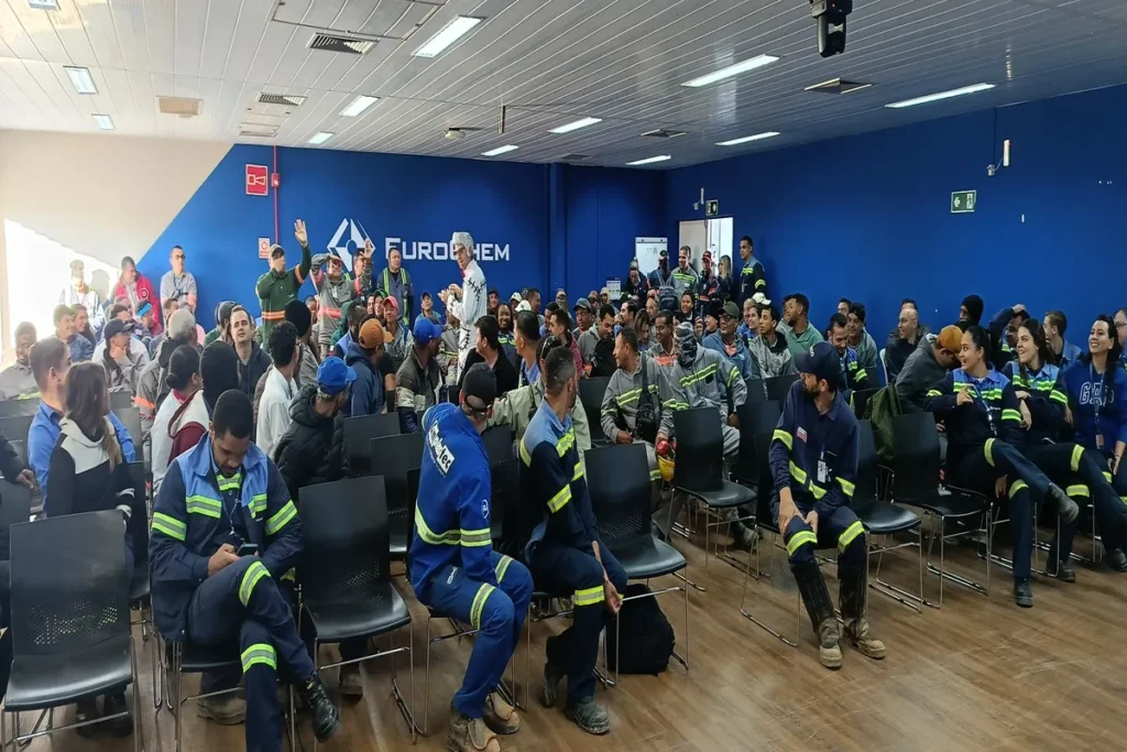 Multidão de funcionários, muitos vestindo uniformes de trabalho, sentados em cadeiras em um auditório azul, assistindo a uma palestra sobre dor lombar crônica.