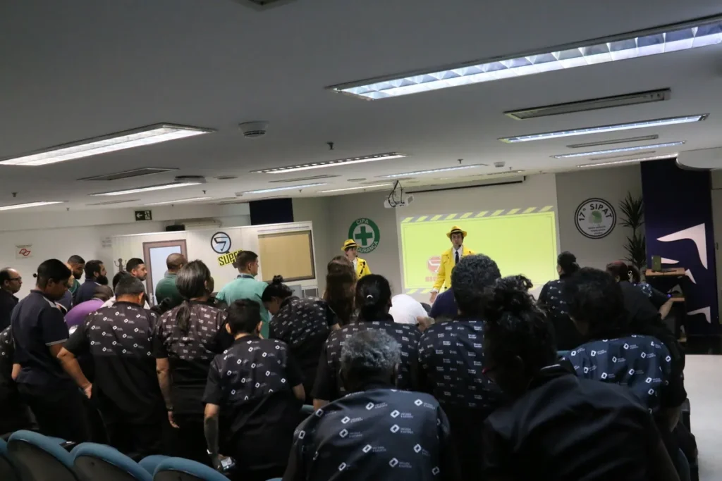 Um palestrante vestido com um uniforme de segurança amarelo e chapéu está em um palco, conduzindo uma palestra em um evento de segurança, provavelmente a SIPAT. Ele está acompanhado por outro profissional no palco. A plateia, em primeiro plano, está de costas, vestindo camisetas escuras padronizadas, atenta à apresentação. No fundo, um projetor exibe uma tela amarela com um símbolo de segurança e o logo da CIPA/SIPAT, destacando a temática de engenheiro de segurança do trabalho e prevenção.
