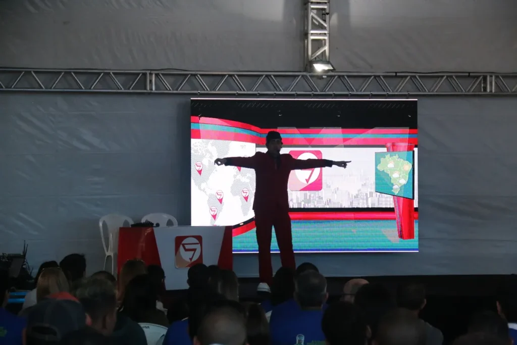 Palestrante motivacional de terno vermelho em pose dramática no palco, com os braços estendidos, em frente a um telão de LED exibindo um mapa-múndi. Palestra focada em Segurança do Trabalho, direcionada a Engenheiros de Segurança e Técnicos de Segurança.